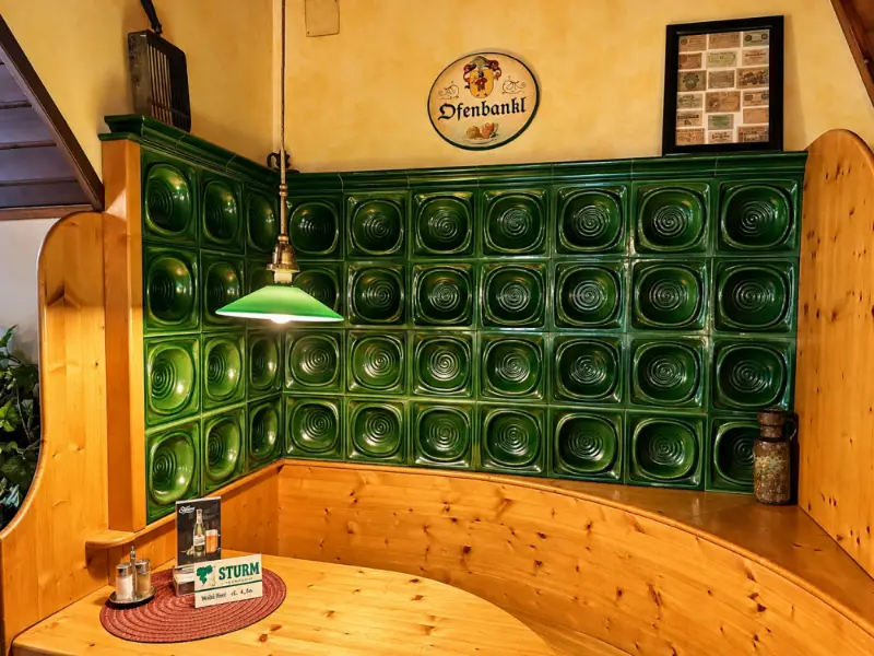 Grüner Kachelofen mit Eckbank, darüber das Schild „Ofenbankl“